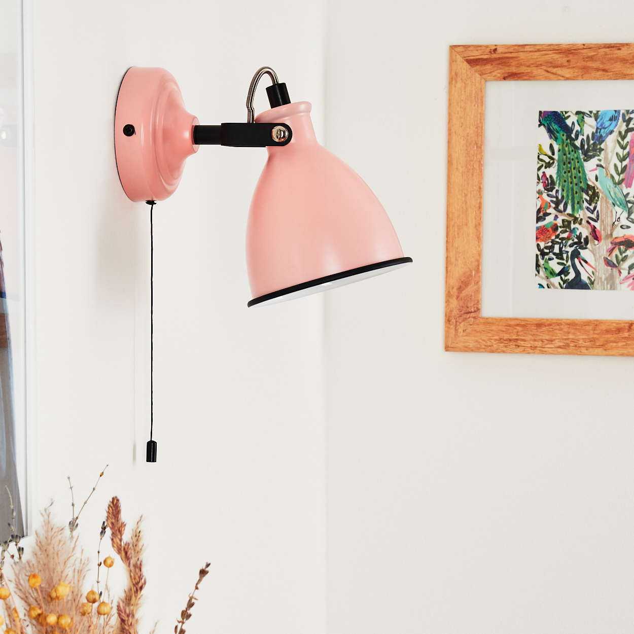 Eine rosafarbene Wandlampe mit Zugschnur ist an einer weißen Wand neben einem gerahmten Bild montiert.