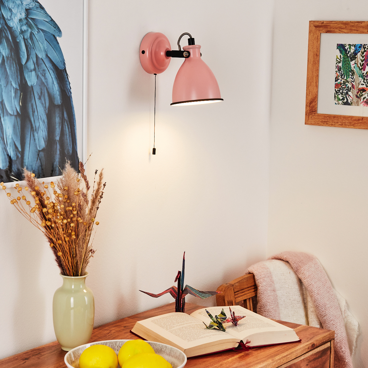 Ein rosafarbener Wandleuchter beleuchtet einen Schreibtisch mit Büchern, Blumen und Zitronen.