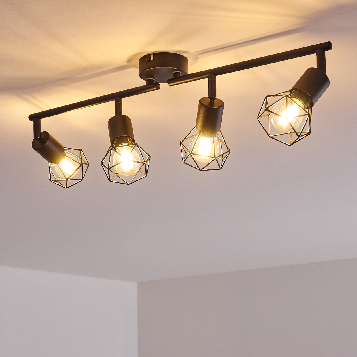 Schwarzer Deckenstrahler mit vier Lampen mit geometrischen Drahtschirmen und sichtbaren Glühbirnen.