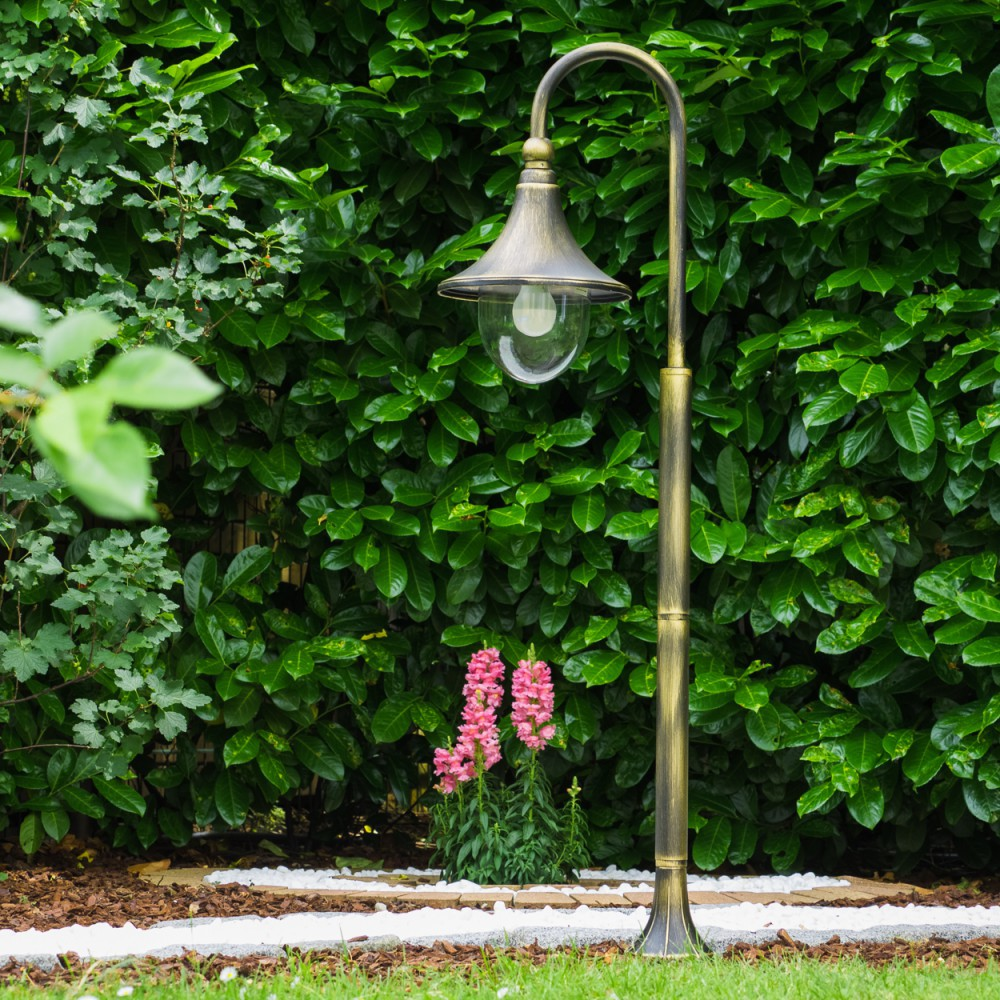Ein bronzener Gartenlichtmast steht vor einer dichten grünen Hecke mit rosa Blumen.