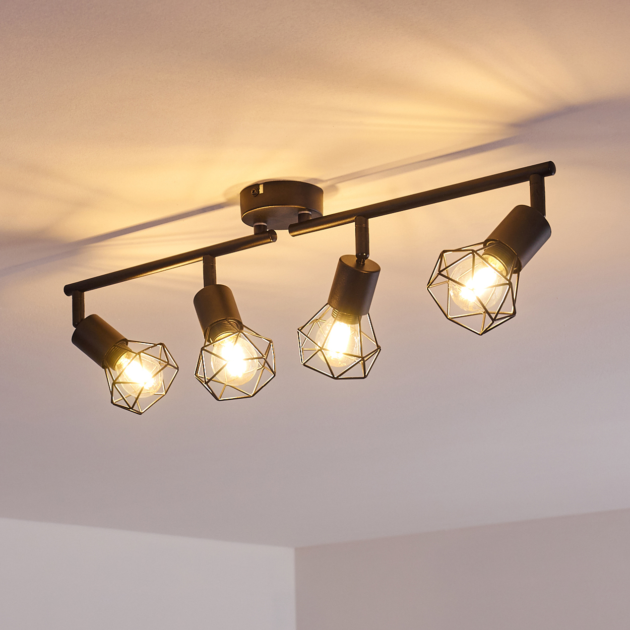Schwarzer Deckenstrahler mit vier Lichtern und Drahtkäfigschirmen, der die Decke beleuchtet.