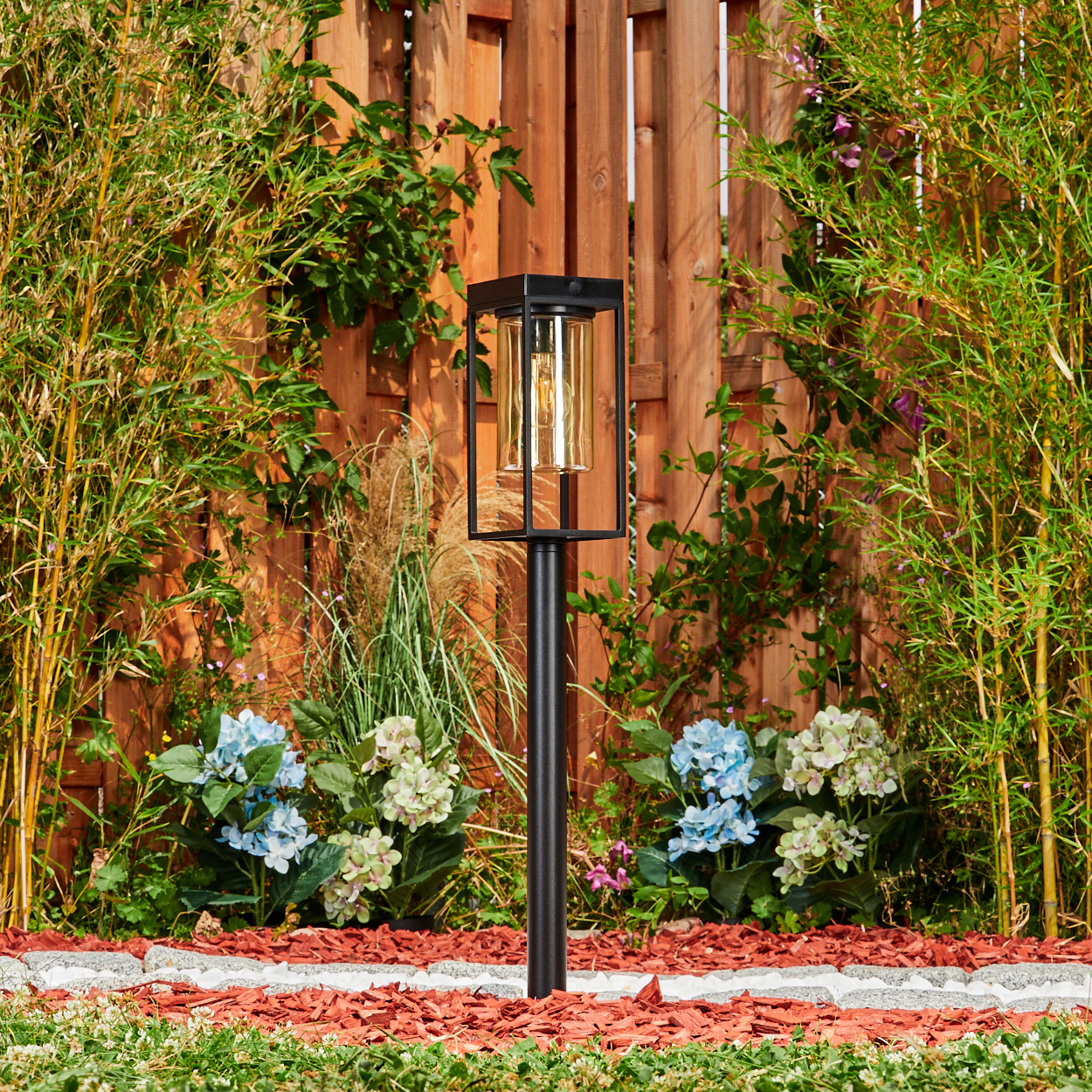 Moderner Gartenpfosten mit Laterne, umgeben von Bambus, Blumen und Holzlattenzaun.