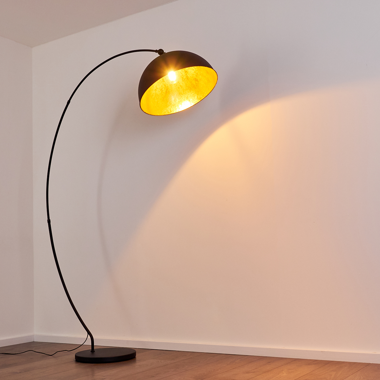 Ein schwarzer Bogenlampenschirm mit goldener Innenseite beleuchtet eine weiße Wand.