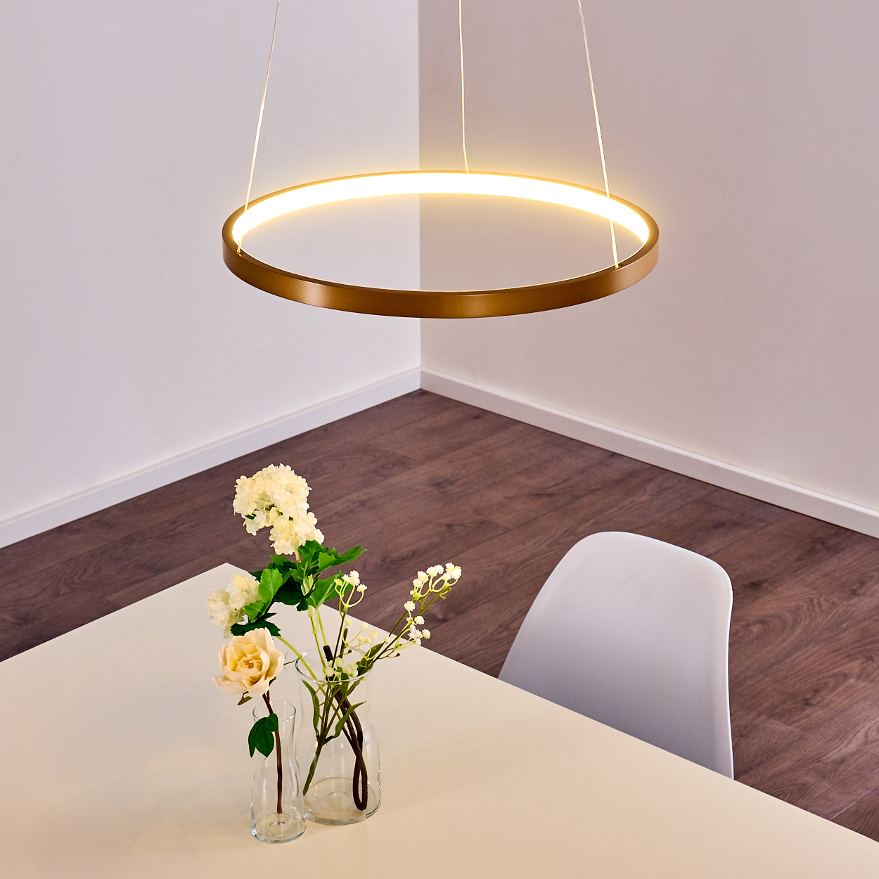 Ein runder goldener Pendelleuchter hängt über einem weißen Tisch mit Blumen in Vasen und einem weißen Stuhl.