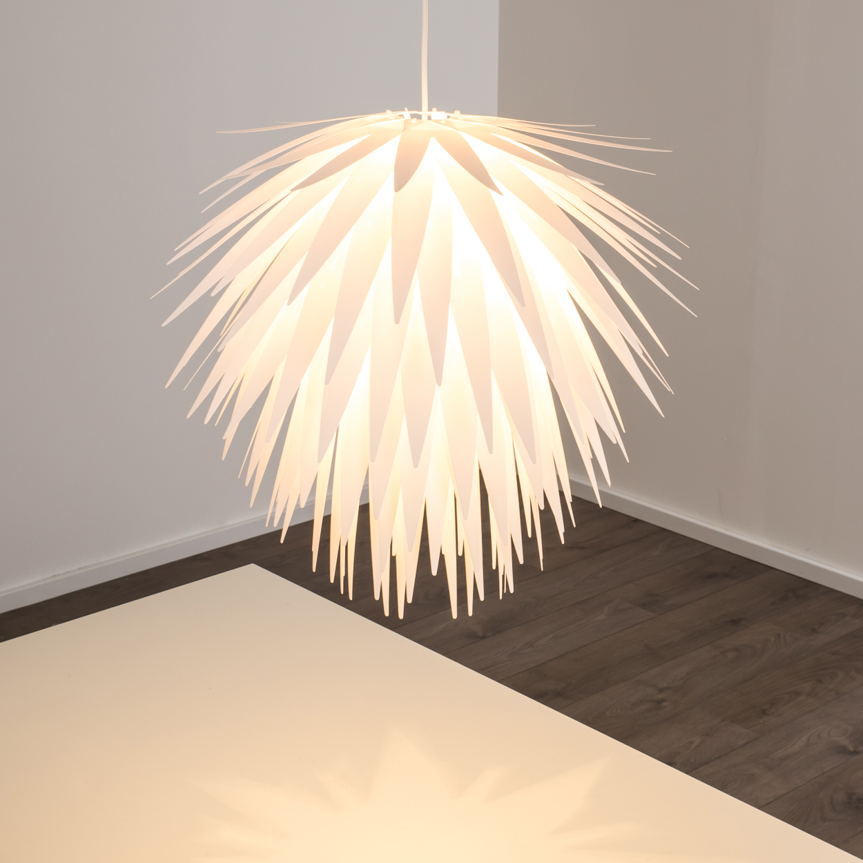 Eine weiße Lampe mit spitzen Zacken hängt über einem weißen Tisch und dunklem Holzboden.