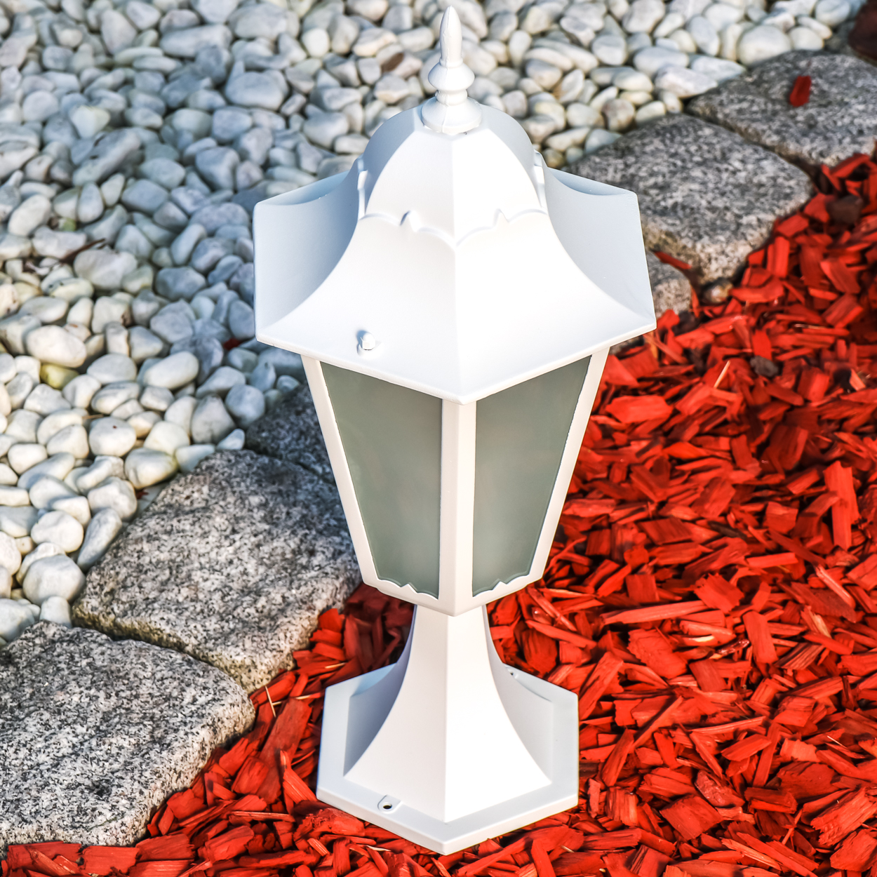 Ein weier Gartenlichtpfosten steht auf rotem Mulch, umgeben von weien Kieselsteinen und grauen Steinen.