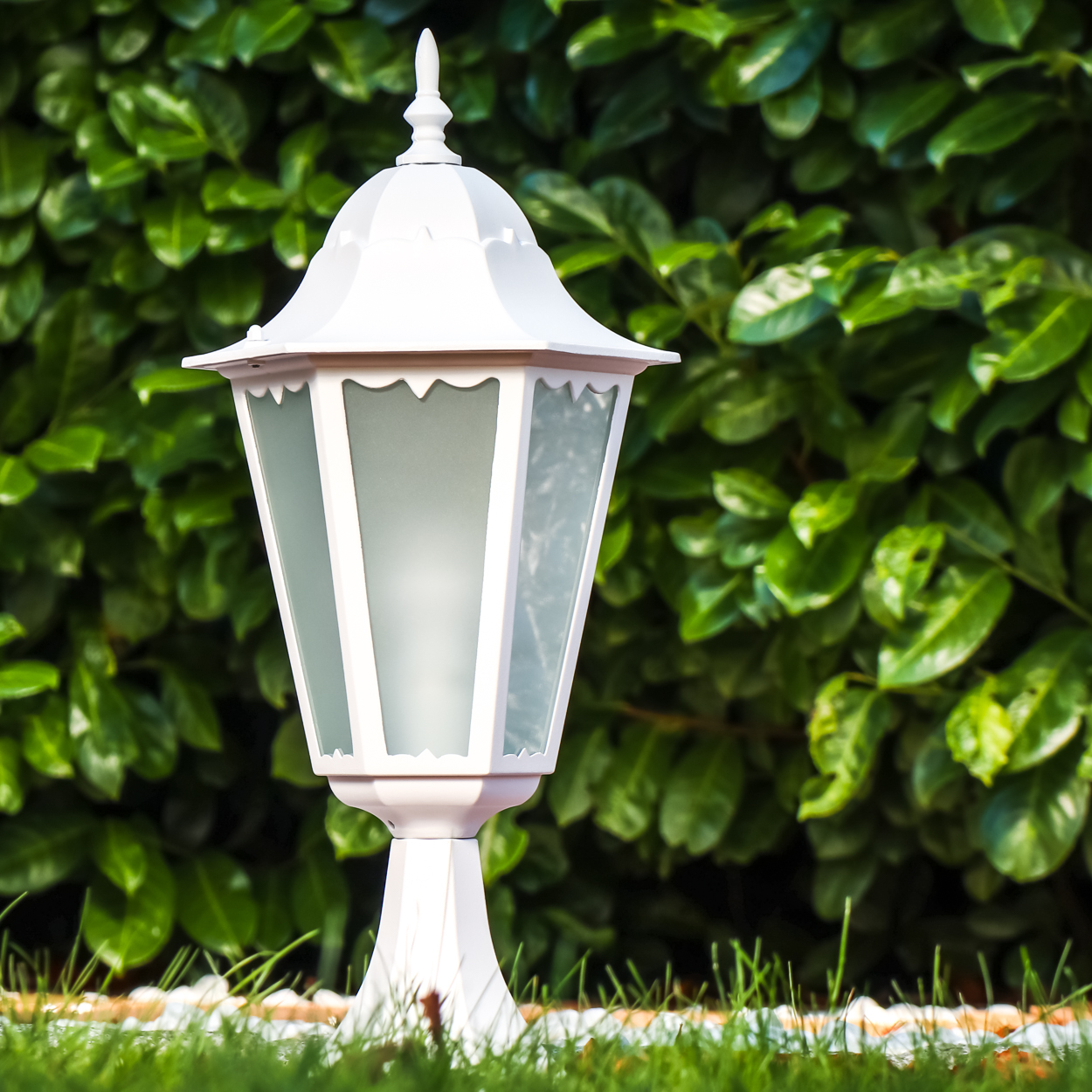 Eine weiße Außenlampe steht im Gras vor einem grünen Busch.