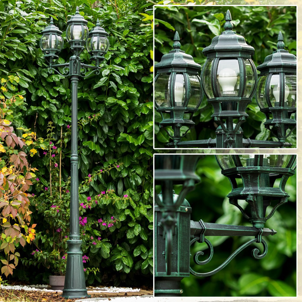 Ein dunkler Laternenpfahl mit drei Lampen steht vor üppigem Grün.