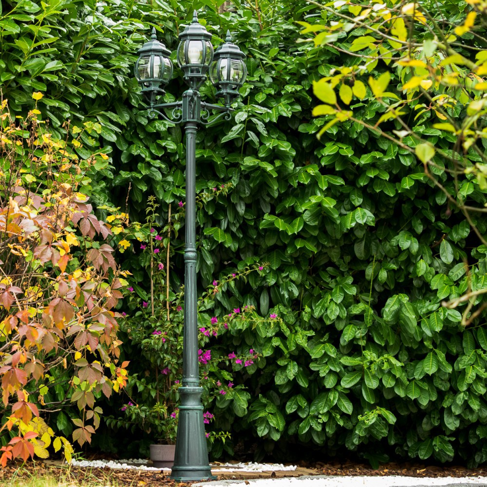 Ein dunkler Laternenpfahl mit drei Lampen steht vor einer dichten grünen Hecke.