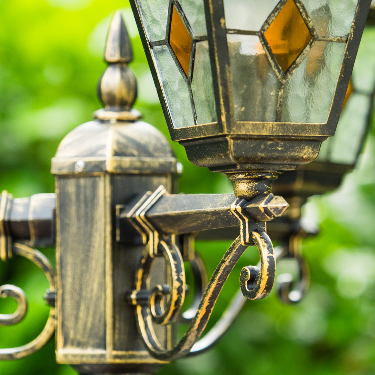 Ein goldener Laternenpfahl mit bunten Glasfenstern vor grünem Laub.