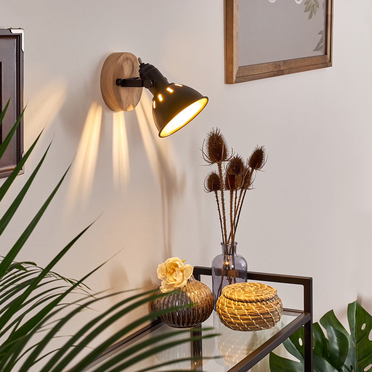 Ein schwarzer Wandspot mit Holzsockel beleuchtet die Wand. Davor stehen Deko-Objekte.