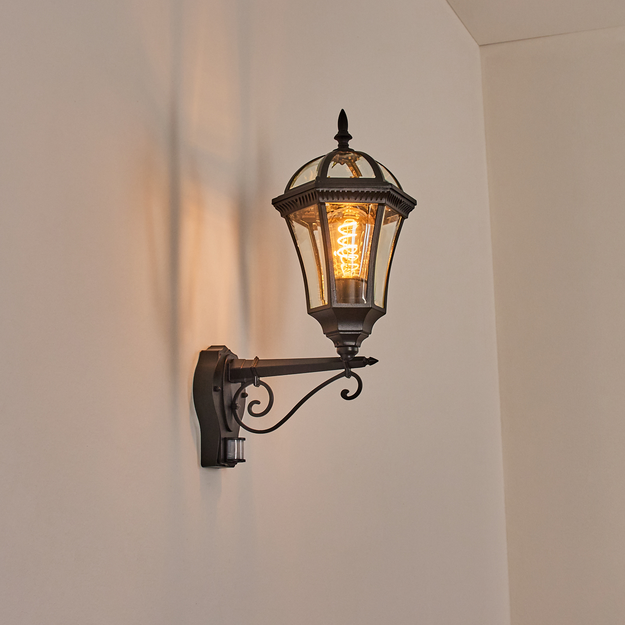 Eine dunkle Wandlampe im Laternenstil mit einer Glühbirne leuchtet auf einer cremefarbenen Wand.