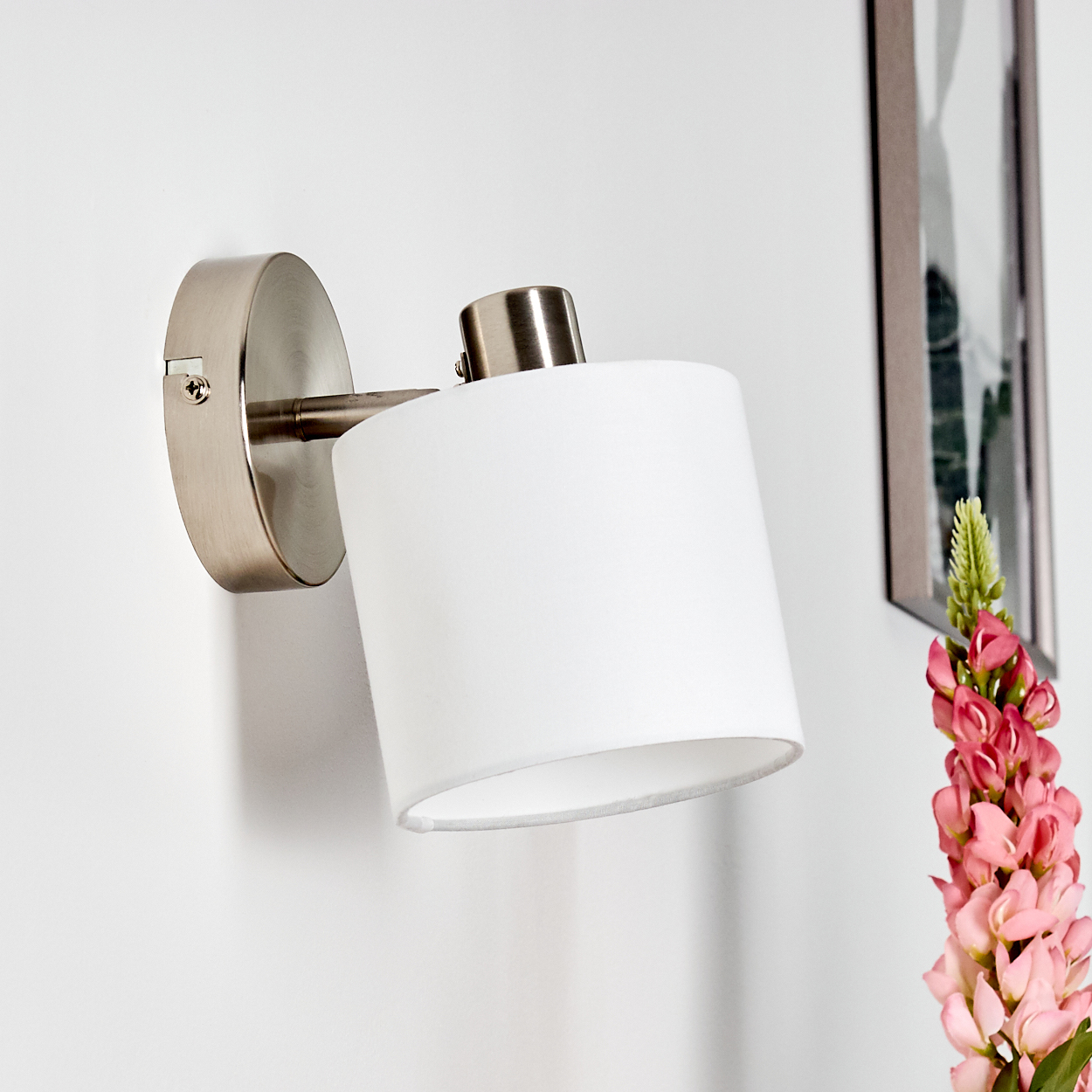 Eine weiße Wandleuchte mit Metallarm hängt an der Wand neben einem Bild und Blumen.