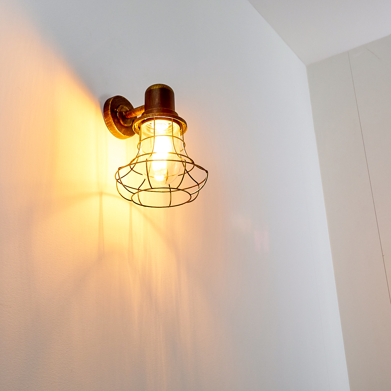 Eine Wandleuchte mit Käfigschirm leuchtet warmes Licht an eine weiße Wand.