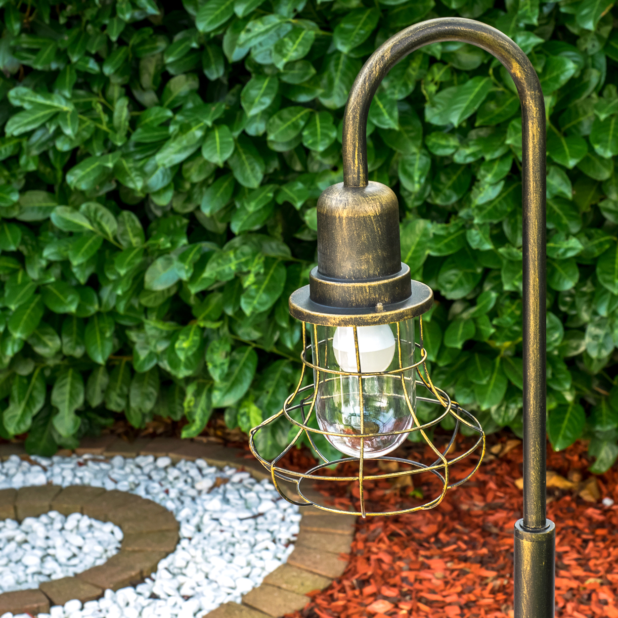 Eine rustikale Lampe mit Glaskolben und Drahtkorb steht vor üppigem Grün.