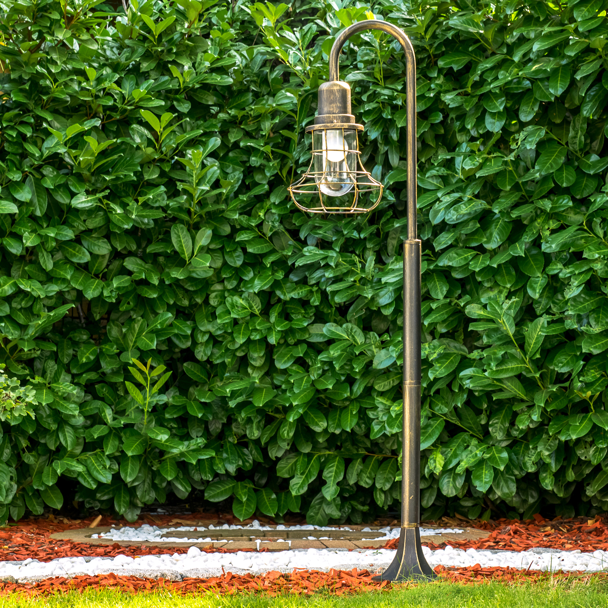 Ein goldener Laternenpfahl steht vor einer dichten grünen Hecke.