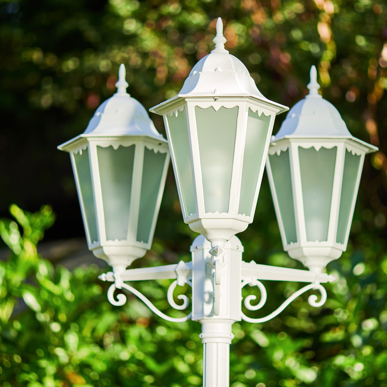 Weiße dreiflammige Lampe im Freien mit grünen Bäumen im Hintergrund.