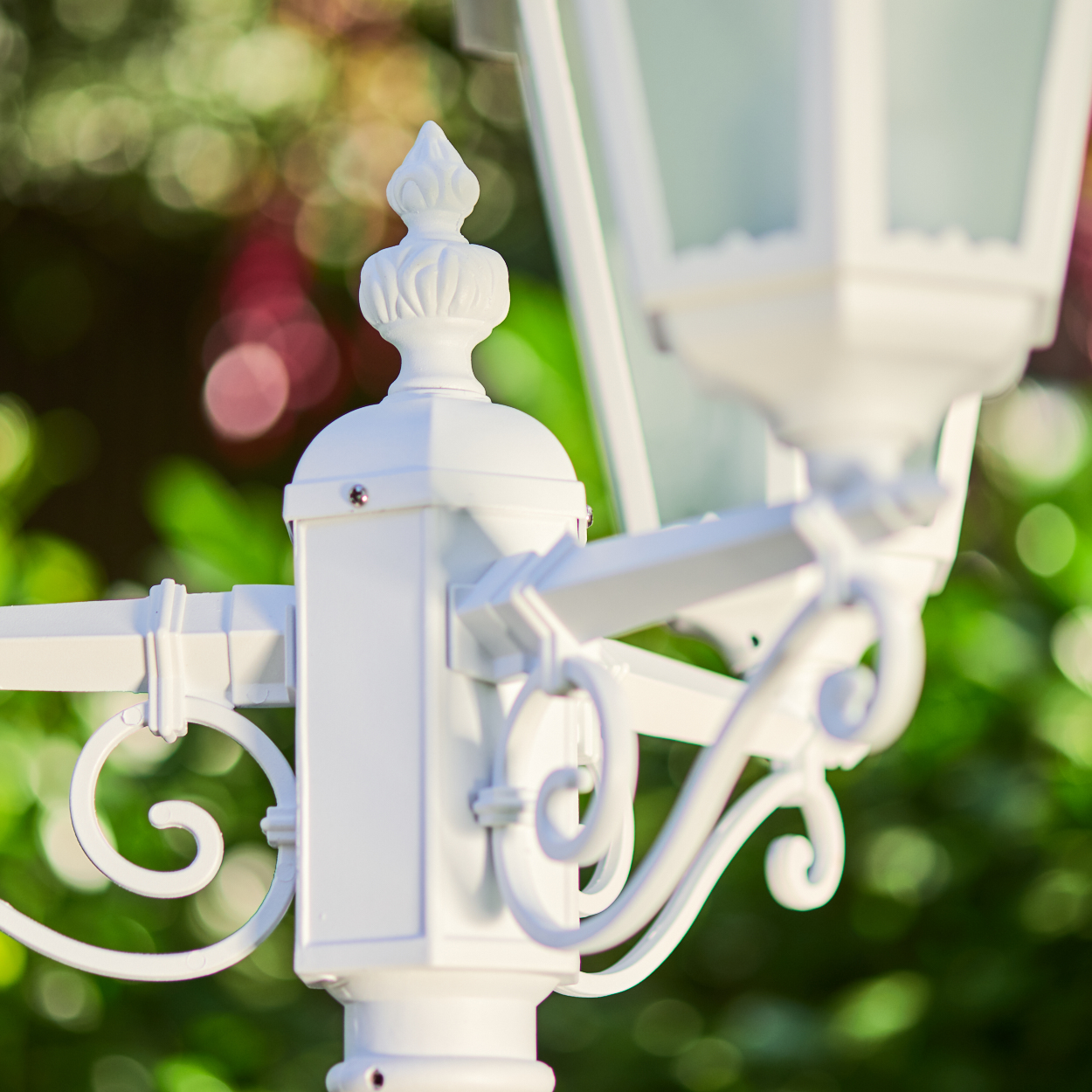 Eine weiße schmiedeeiserne Laterne mit geschwungenen Verzierungen im Vordergrund, Bokeh-Hintergrund.