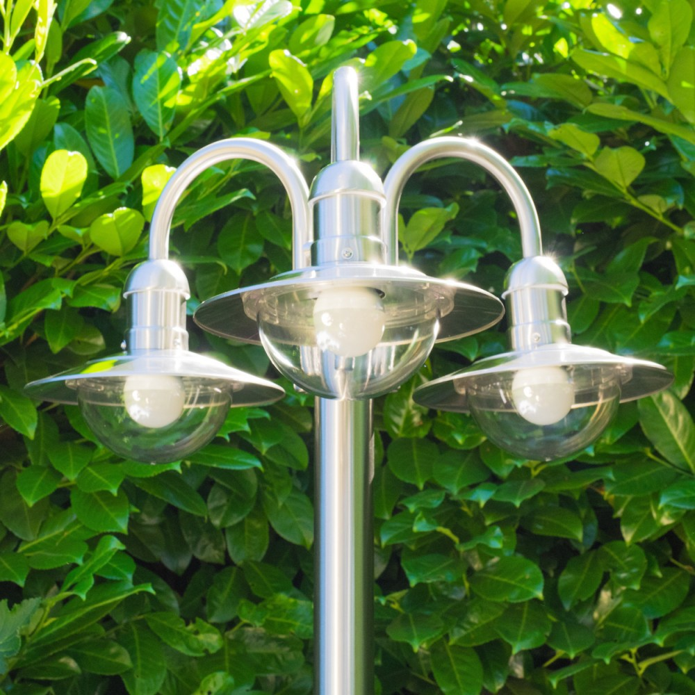 Ein silberner Laternenmast mit drei Lampen steht vor einem dichten grünen Blätterhintergrund.