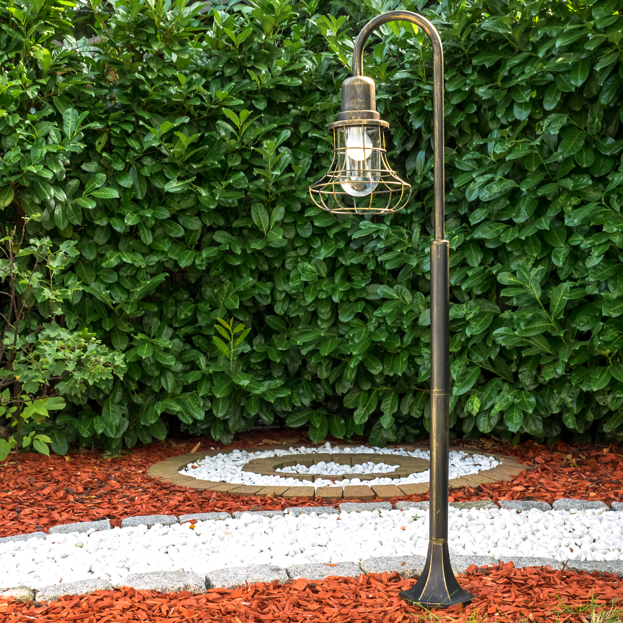 Gartenlampe vor einer grünen Hecke mit rotem Rindenmulch und weißen Steinen.