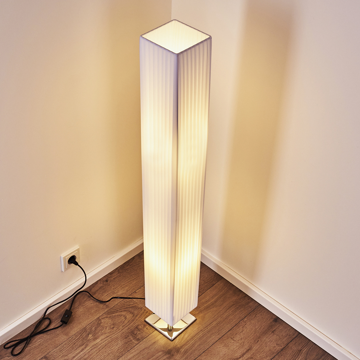 Eine weiße Stehlampe mit Faltenstoff steht in einer Ecke, beleuchtet den Raum.