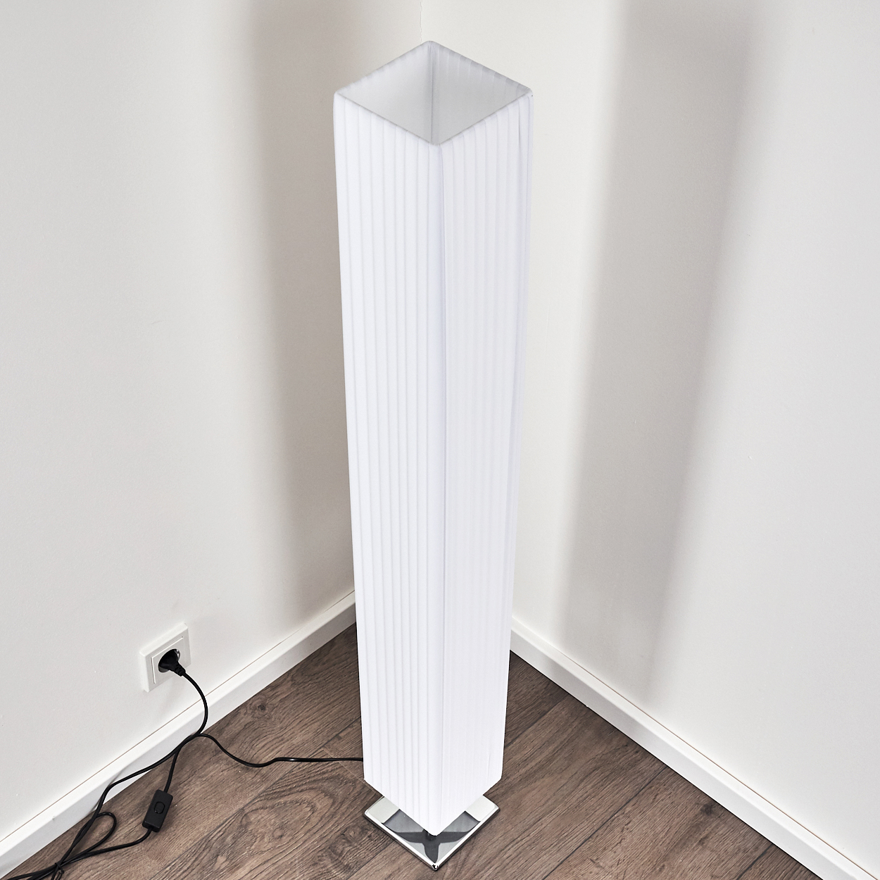 Eine weiße Stehlampe mit plissiertem Lampenschirm steht in einer Ecke auf Holzboden.