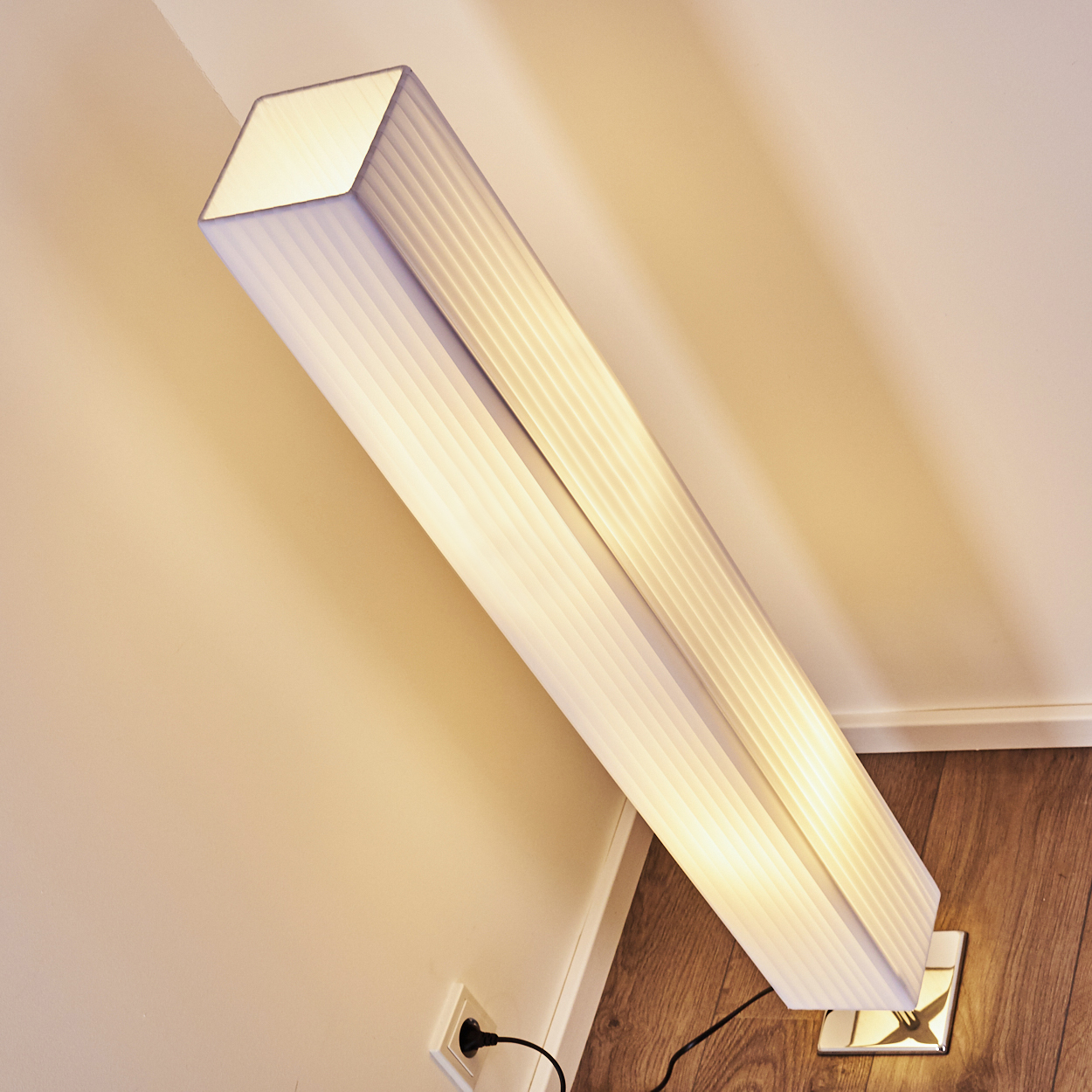 Eine moderne Stehlampe mit weißem Stoffschirm steht auf einem Holzboden neben einer Wand.