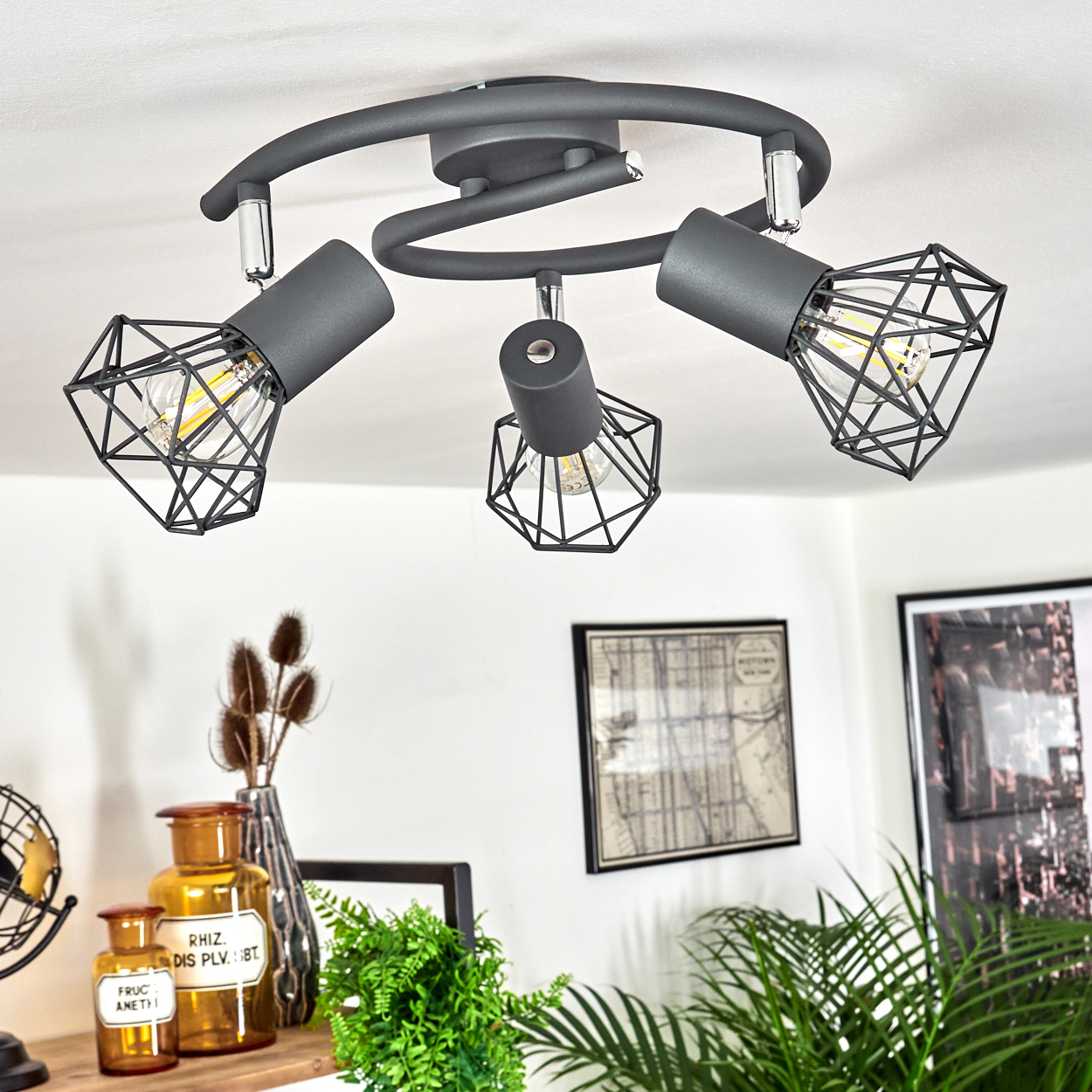 Grauer Deckenstrahler mit drei Lampen und Drahtkäfigschirmen über einem Regal mit Dekoration.