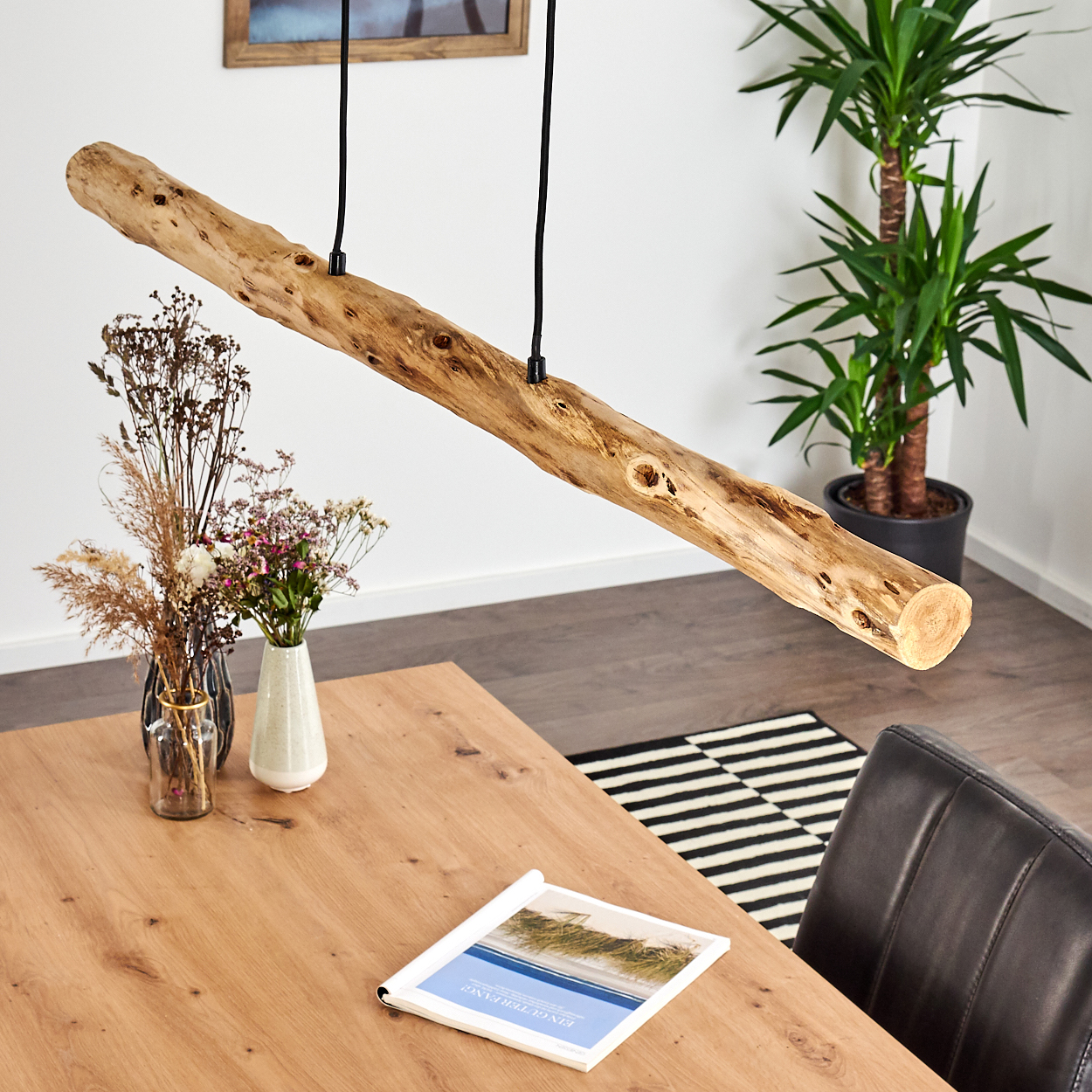 Ein rustikaler Ast als Hängelampe über einem Holztisch mit Blumen und einem Buch.