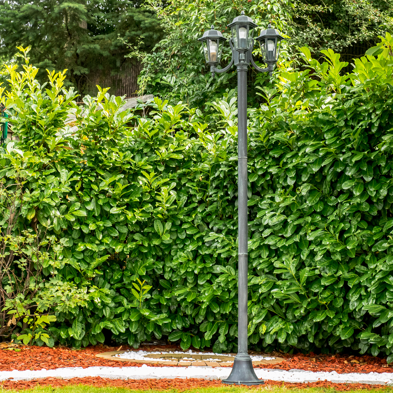 Ein dunkler Laternenpfahl steht vor einer üppigen grünen Hecke.
