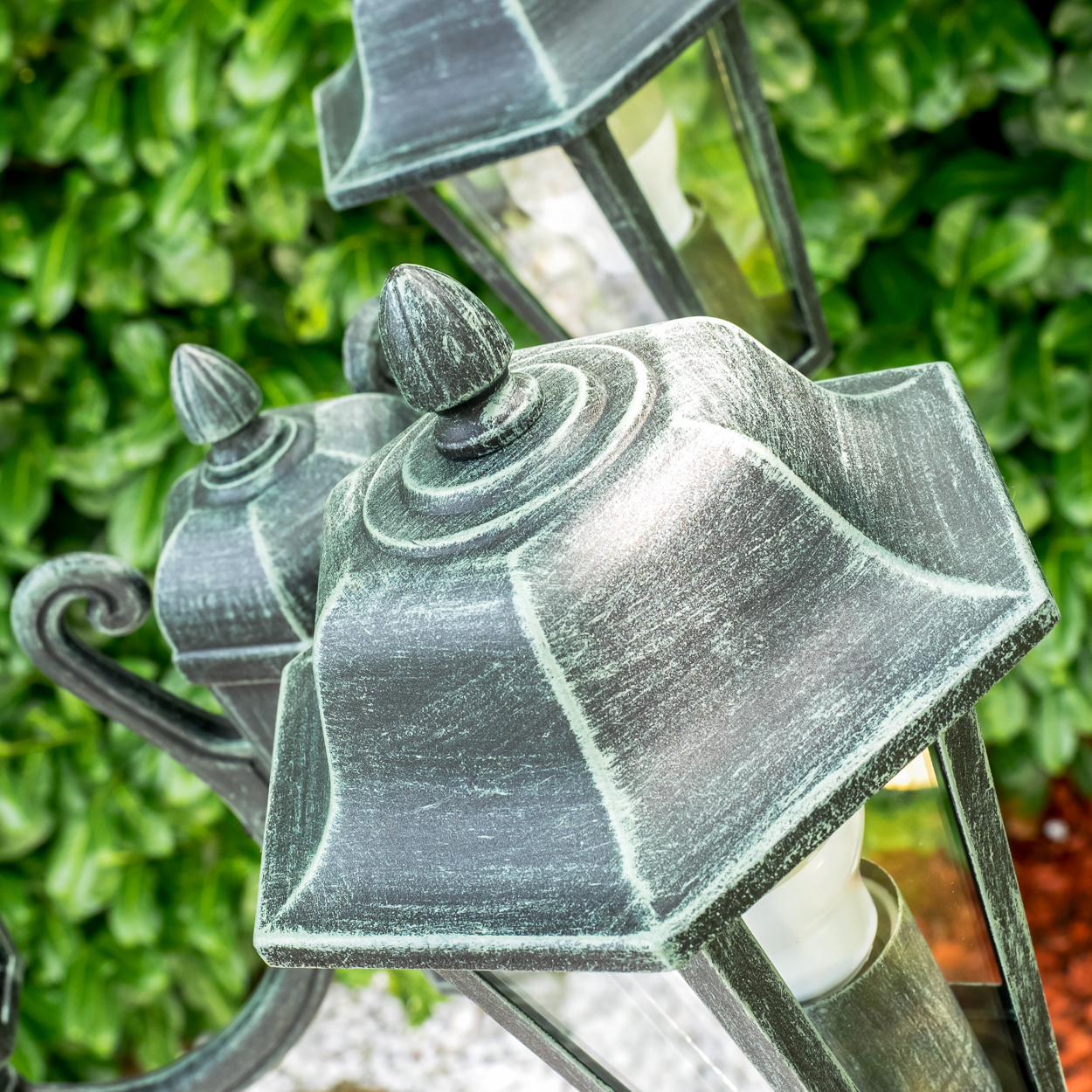 Detailaufnahme von dunklen Laternen mit patinaartiger Patina und grüner Hecke im Hintergrund.