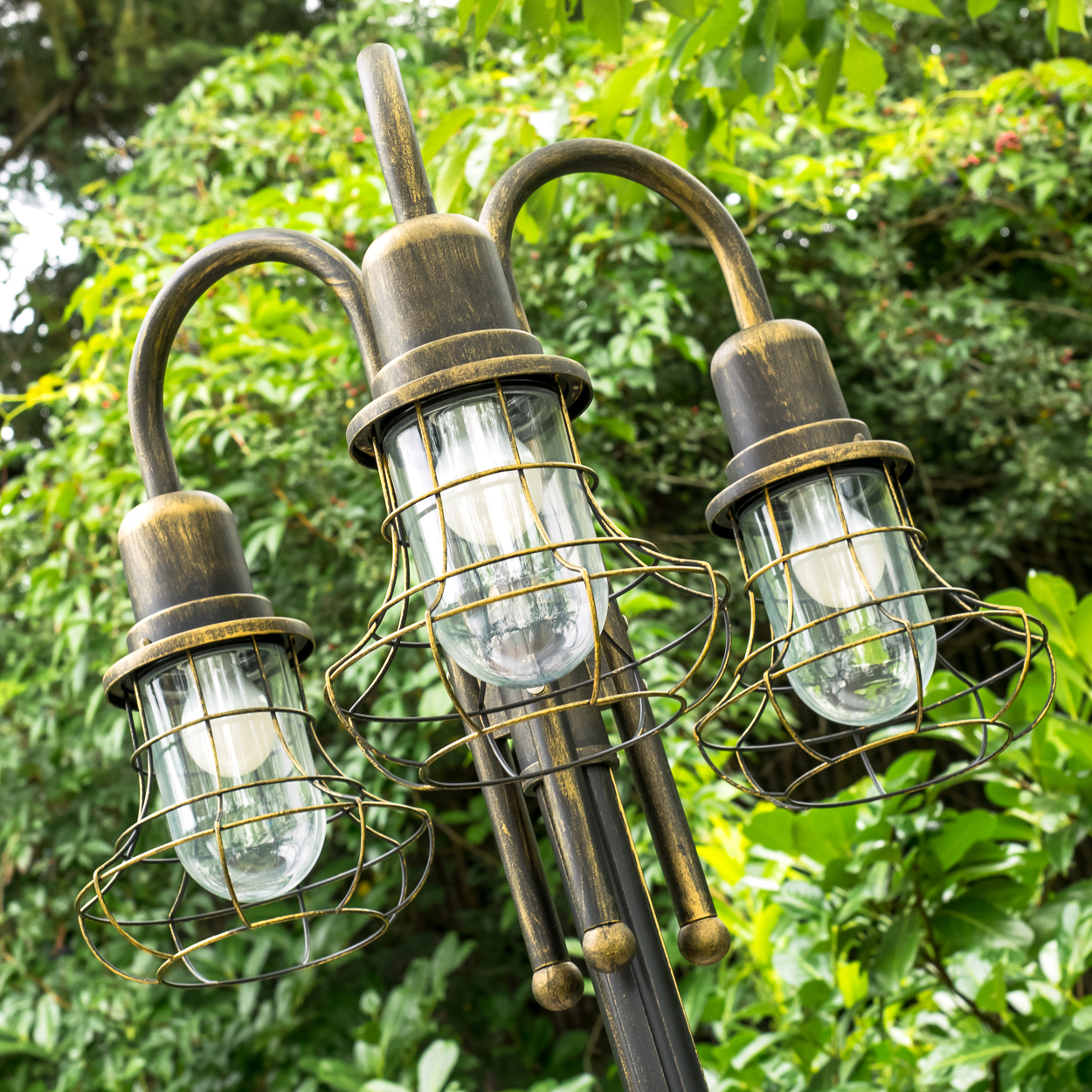 Eine bronzefarbene Lampe mit drei Glasglocken und Drahtschutz vor üppigem Grün.
