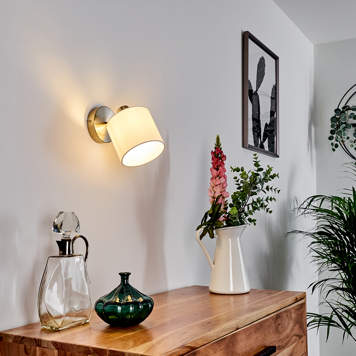 Ein Wandlicht beleuchtet eine weiße Wand über einer Holzkommode mit Dekorationen.