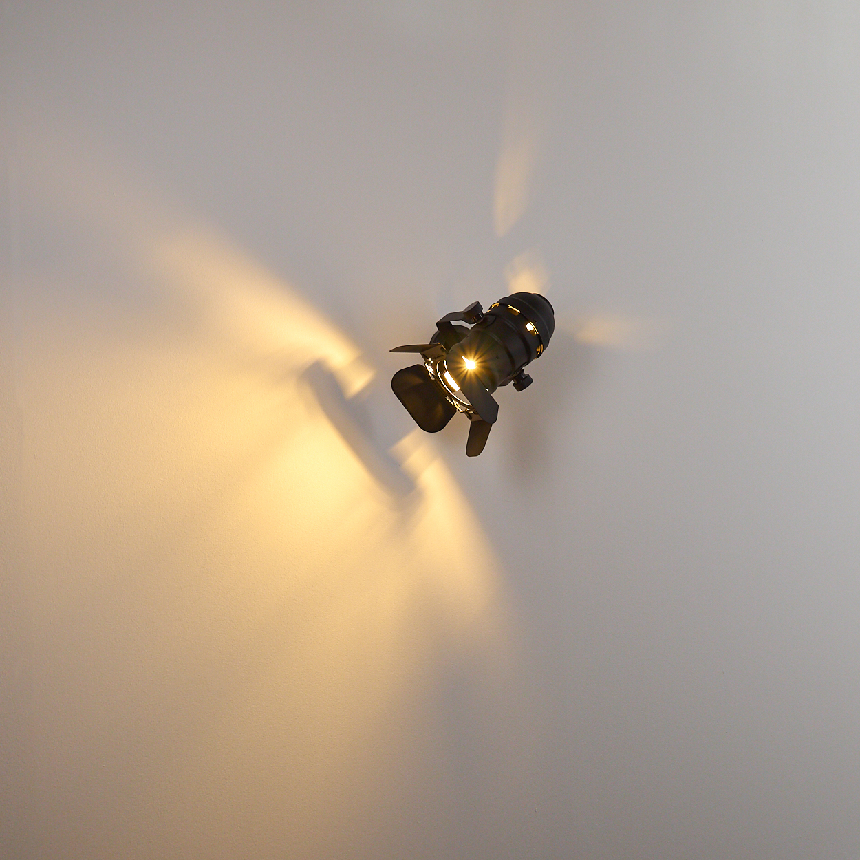 Ein schwarzer Scheinwerfer leuchtet auf einer weißen Wand, wirft gelbes Licht und Schatten.