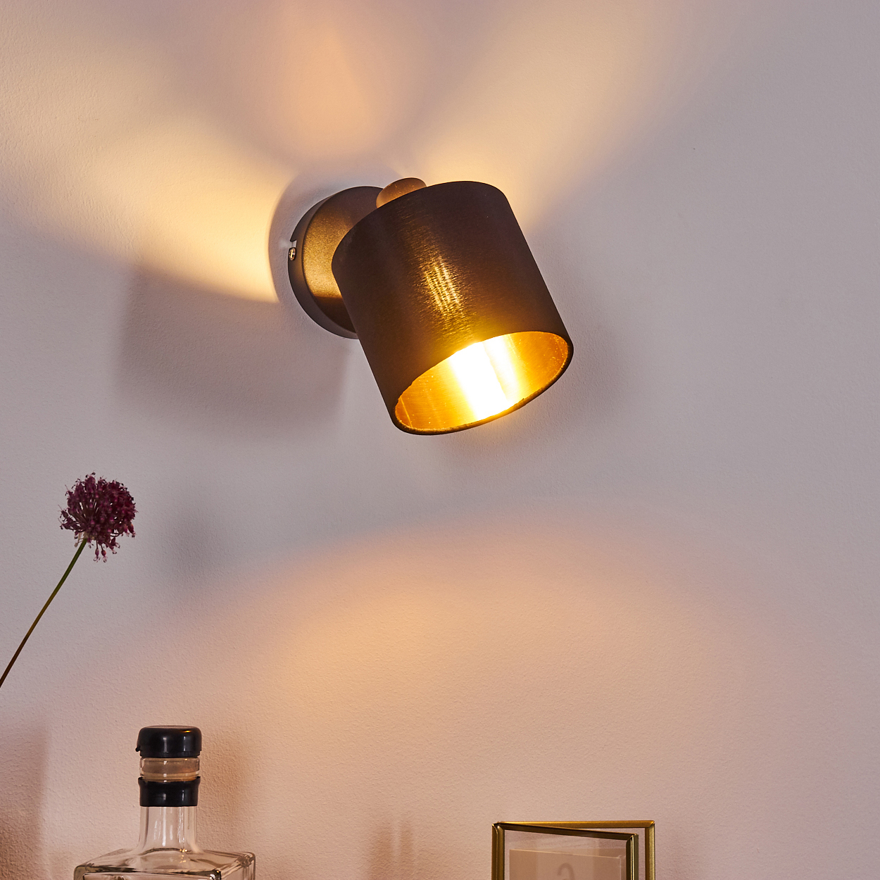 Ein schwarzer Wandstrahler mit goldenem Innenleben beleuchtet die Wand. Daneben eine Blume.