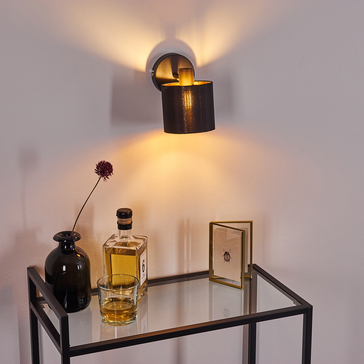 Ein schwarzer Wandleuchter beleuchtet eine Glasablage mit einer Vase, Flasche und einem Bilderrahmen.