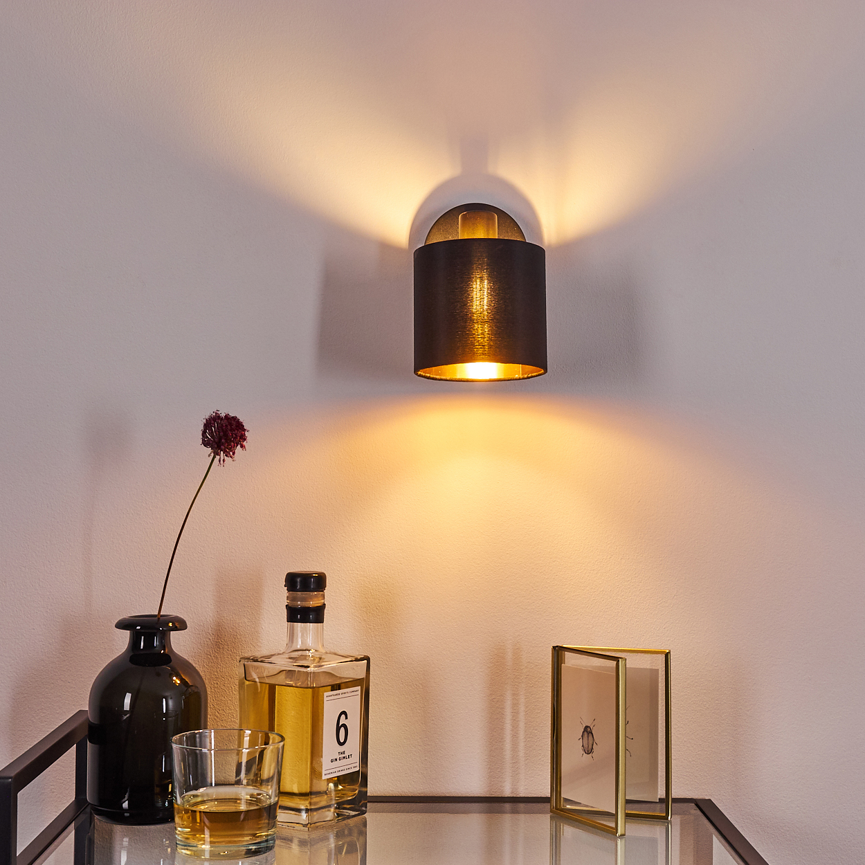Eine Wandleuchte beleuchtet eine Wand, daneben stehen eine schwarze Vase, ein Glas und eine Flasche.