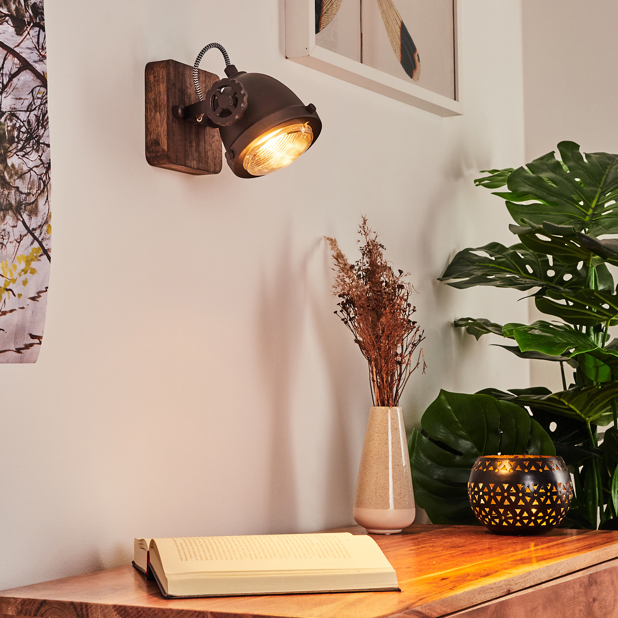 Ein Wandstrahler beleuchtet ein offenes Buch auf einem Holzmöbel neben einer Vase mit Trockenblumen und einer Pflanze.