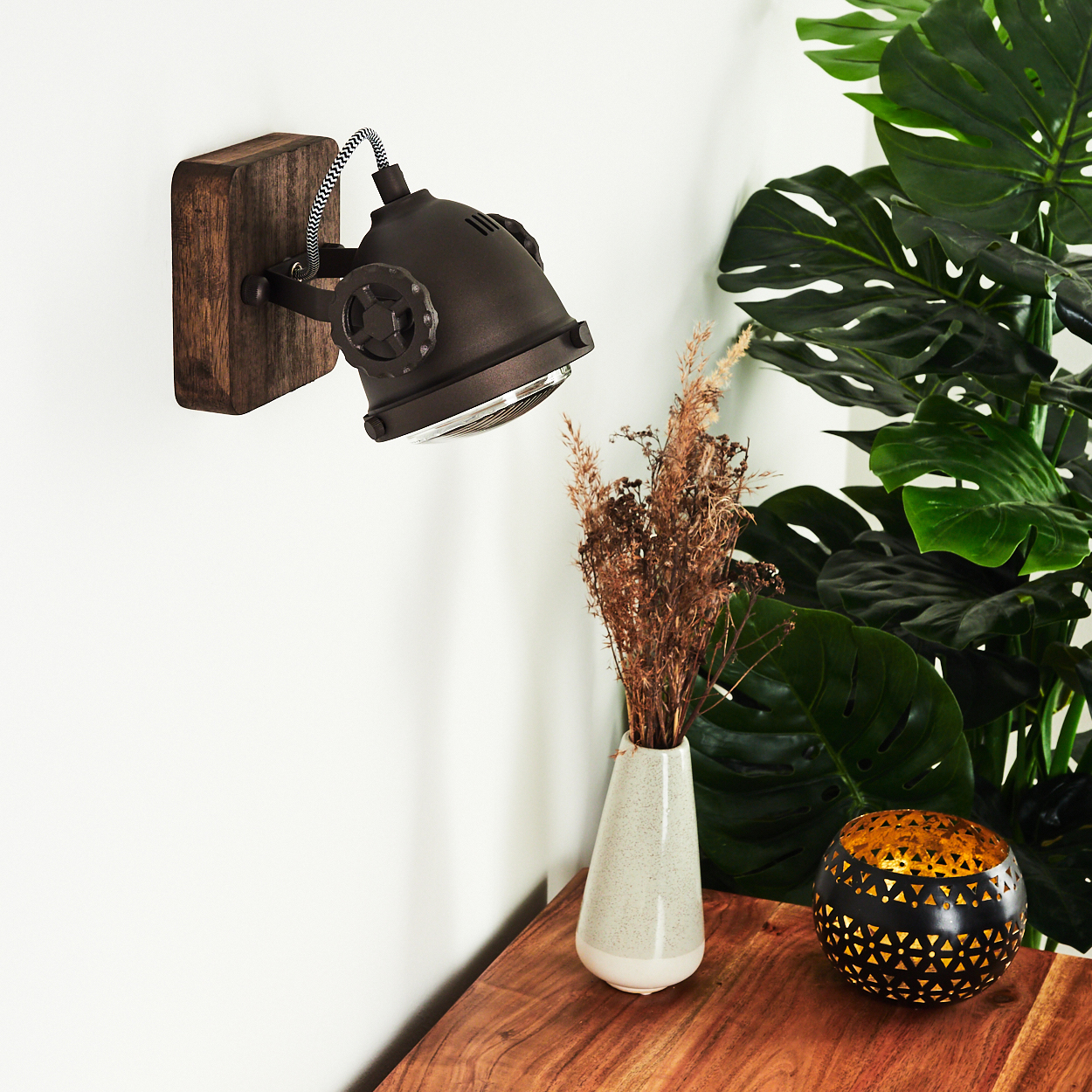 Ein dunkler Wandspot über einem Holztisch mit Vase, Trockenblumen und Laterne.