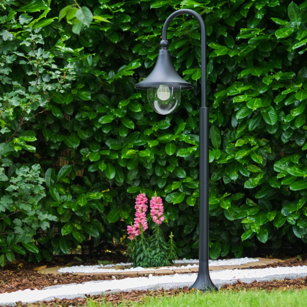 Ein schwarzer Gartenlichtmast steht vor einer dichten Hecke mit rosa Blumen im Vordergrund.