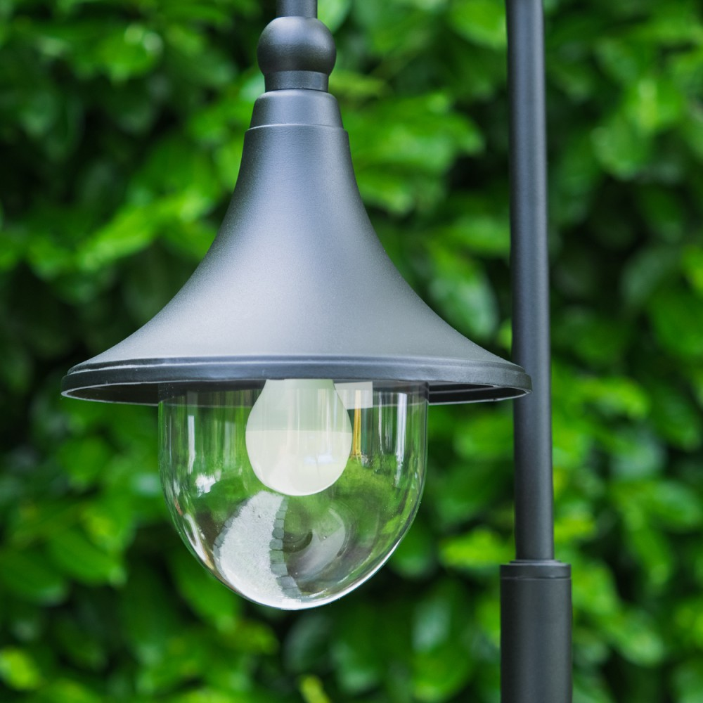 Eine schwarze Außenlampe mit klarem Glasschirm und weißer Glühbirne vor grüner Vegetation.