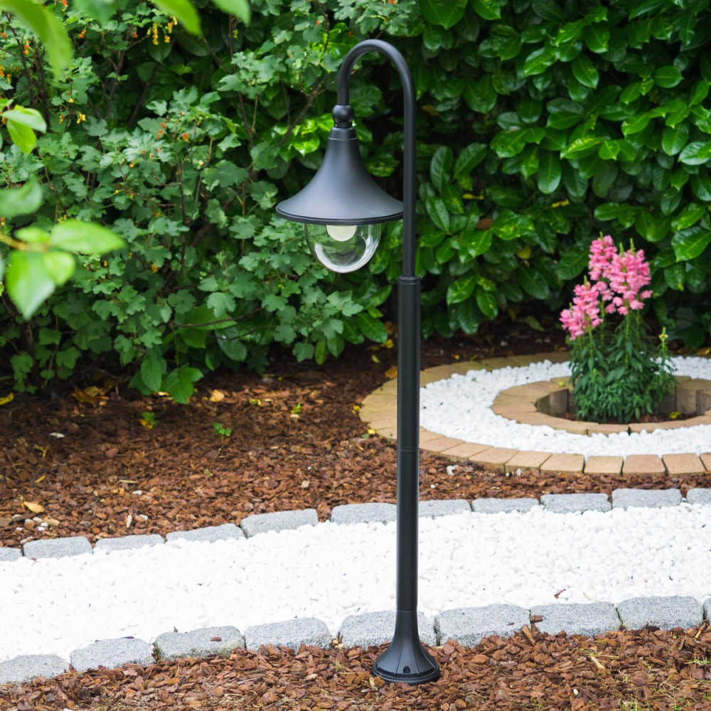 Ein schwarzer Laternenpfahl steht in einem Garten mit weißen Kieseln und grünen Büschen.