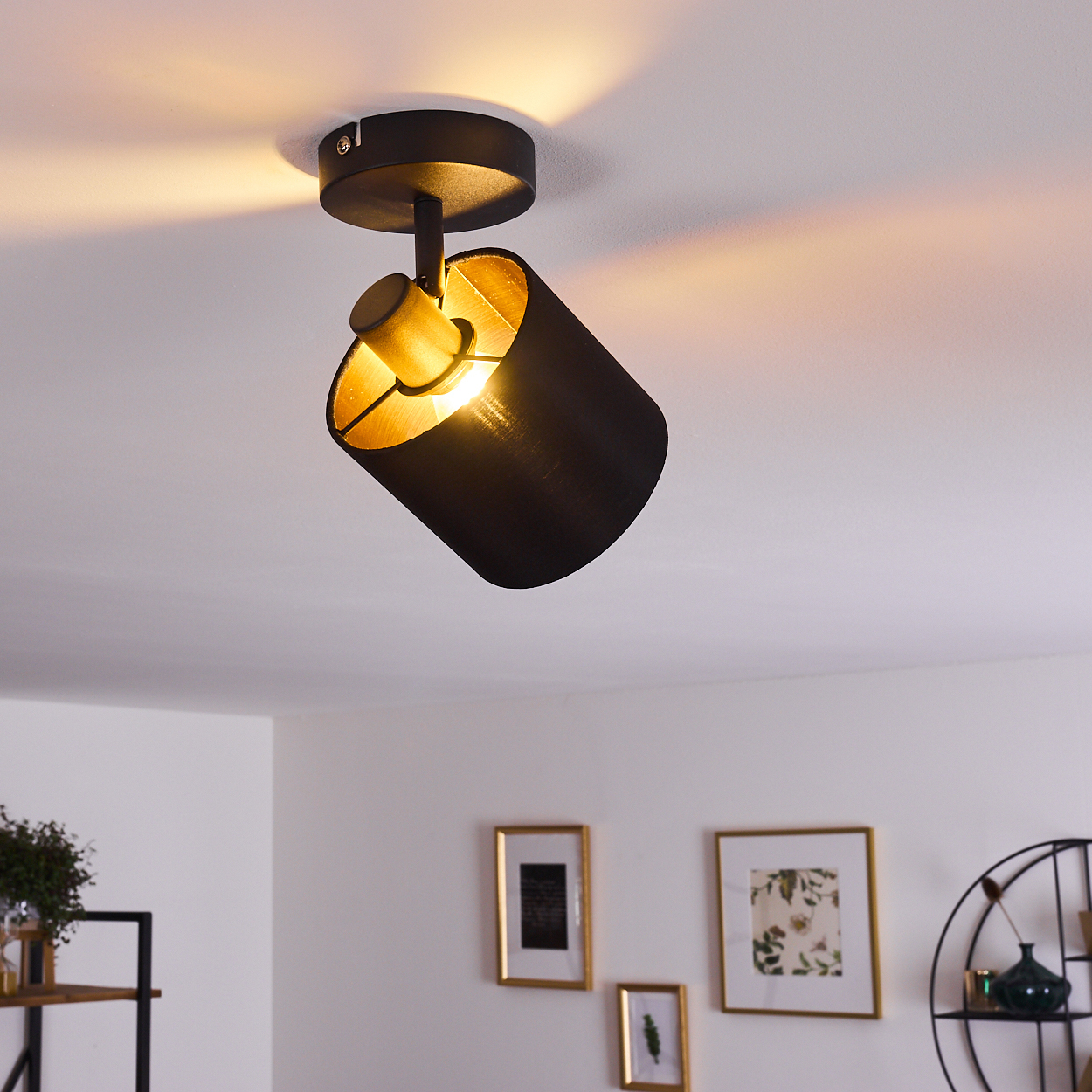 Schwarze Deckenleuchte mit goldenem Innenleben, nach unten geneigt. Warmes Licht beleuchtet die weiße Decke.