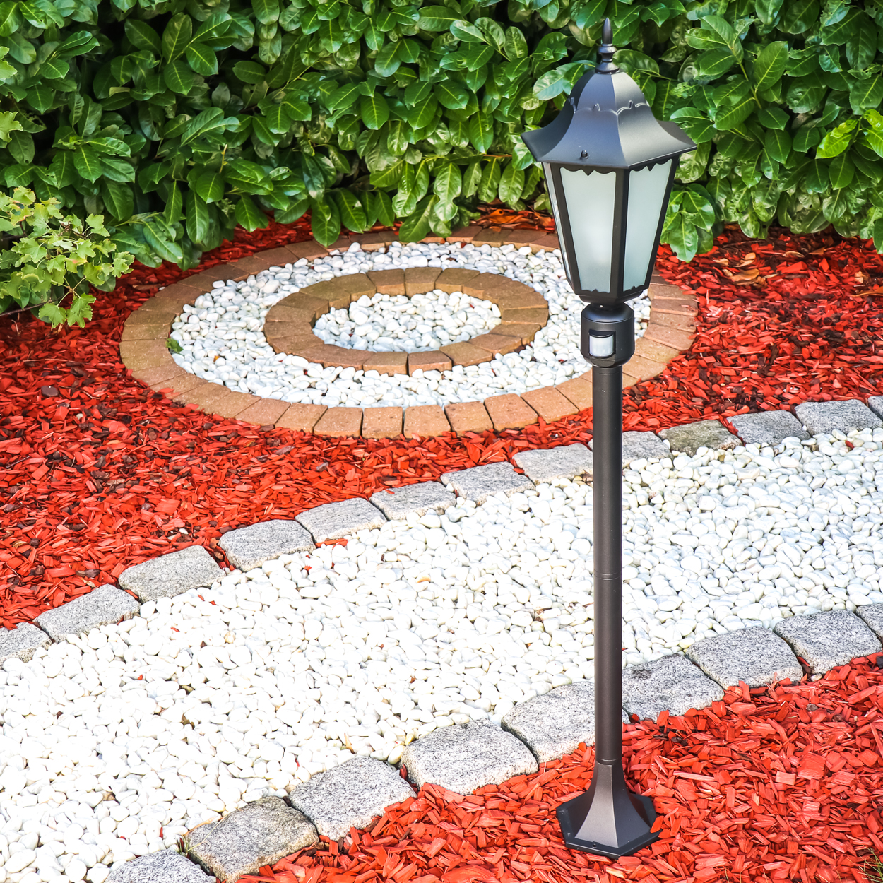Ein schwarzer Laternenpfahl steht auf einem Gartenweg mit weißem Kies und rotem Mulch.