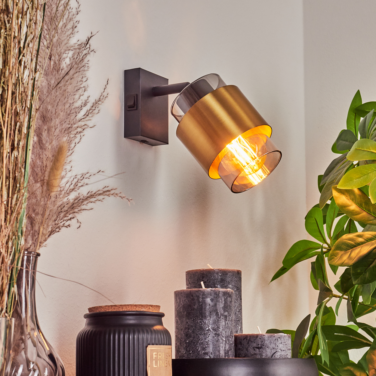 Goldener Wandstrahler mit Glasakzenten, neben einem Strauß getrockneter Gräser und Kerzen.