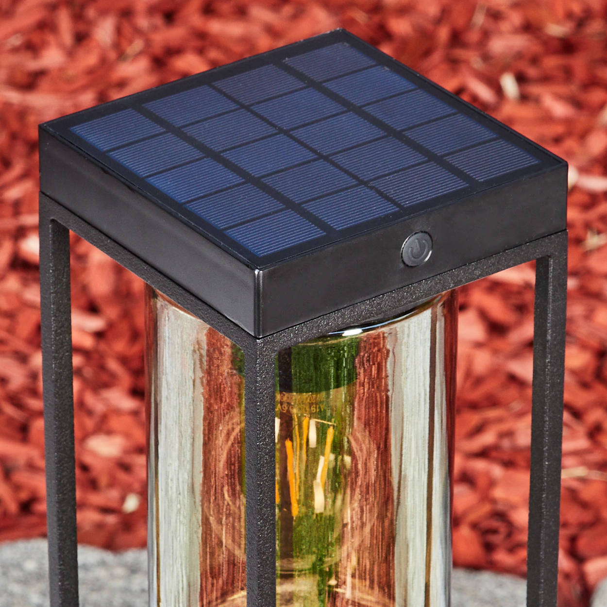 Schwarze Solarlaterne mit Glaszylinder und Glühbirne, auf rotem Mulch.