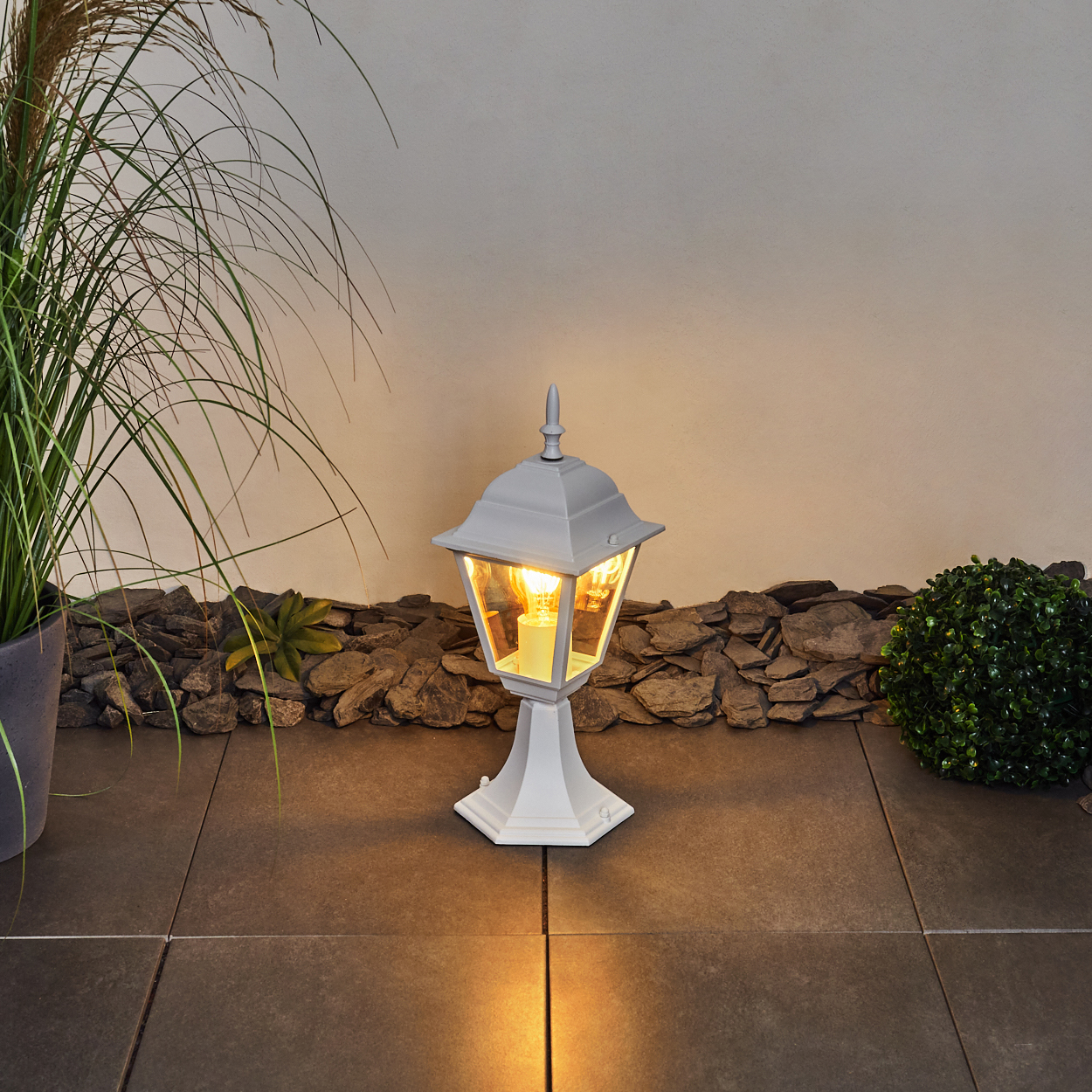 Eine weiße Laterne leuchtet auf einer gefliesten Terrasse mit Pflanzen.