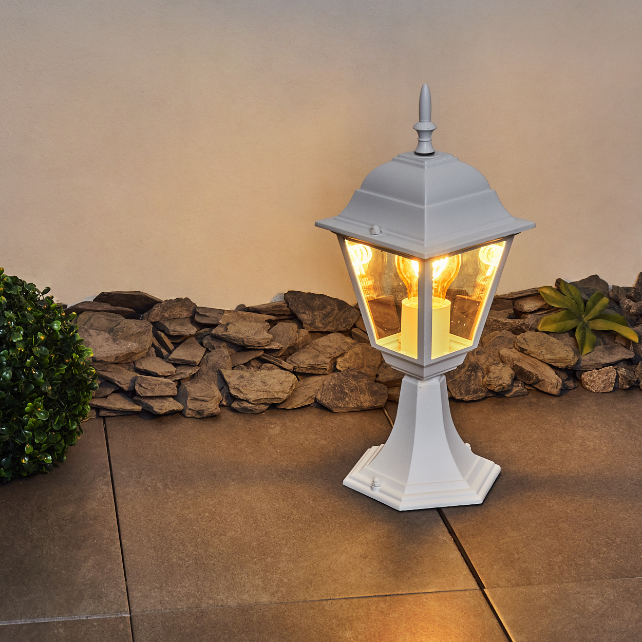 Weiße Außenlampe mit Glühbirnen auf gepflastertem Boden, vor Steinmauer und Büschen.