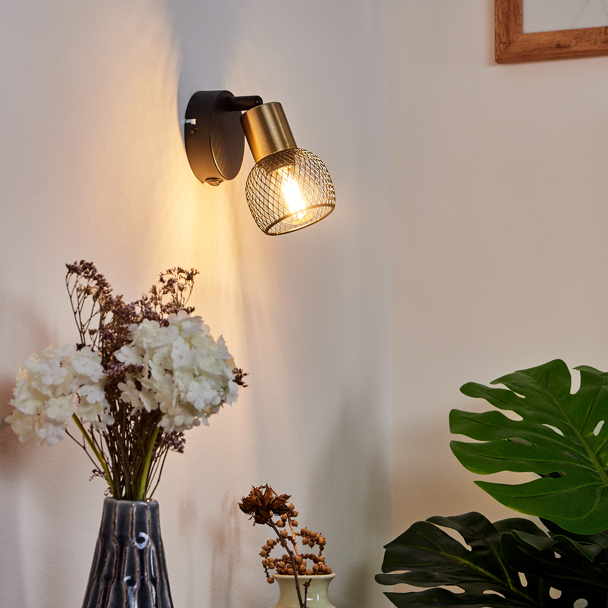 Eine Wandleuchte mit goldenem Käfig-Schirm leuchtet auf eine weiße Wand.