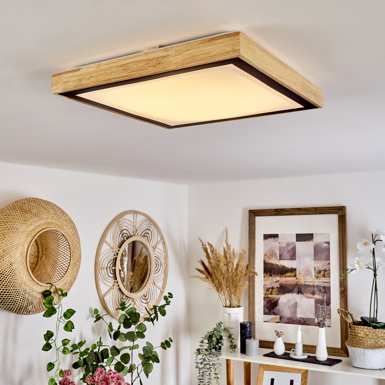Ein quadratisches Deckenlicht mit Holzrahmen hängt über einer Wand, geschmückt mit gewebter Kunst, Pflanzen und einem gerahmten Bild.